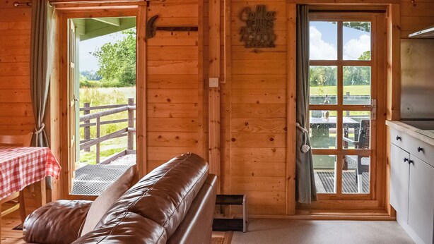 A wooden cabin interior with a leather sofa, a dining table with a red and white checkered tablecloth, and a view of a grassy field through a door. - 1 bedroom accommodation in Berrow, near Malvern (Berrow, near Malvern)