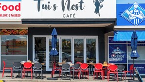 Ein Café mit Außenbestuhlung, darunter rote und graue Stühle sowie ein blauer Sonnenschirm.