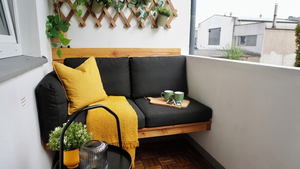 Een balkon met een houten bankje, een tafeltje met een plant en een plantenrek aan de muur.