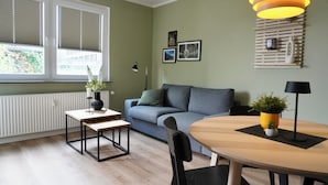 Een moderne woonkamer met een bank, een eettafel en een salontafel. Er zijn twee ramen met zonwering, een lamp en een wandplank met decoratieve objecten.