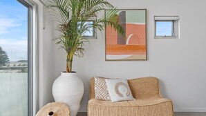 Une pièce dotée d’une grande fenêtre, d’un vase blanc contenant une plante, d’un canapé en osier agrémenté de coussins à motifs et d’un tableau abstrait encadré accroché au mur.