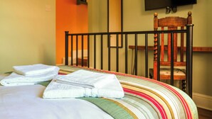 Una cama con sábanas blancas y almohada a rayas, una silla de madera, un televisor montado en la pared y una puerta que da a otra habitación.