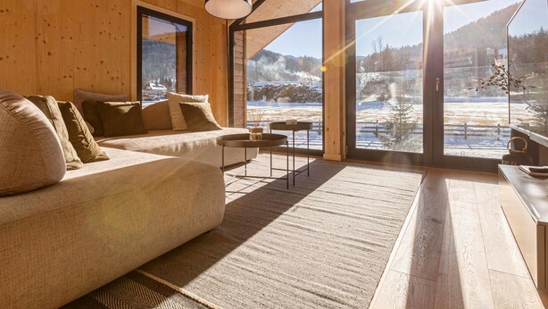 Interior acogedor de una cabaña con sofá, mesa y ventanas amplias que ofrecen vistas a un paisaje nevado.