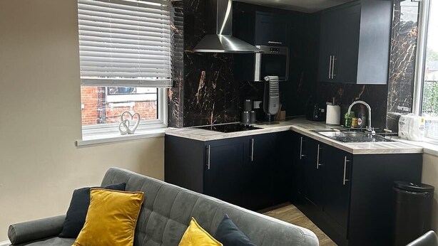 A modern kitchen with dark cabinets, a grey sofa with yellow cushions, and a window with blinds. - Leeds Urban Retreat- Pet Friendly Flat (Leeds)
