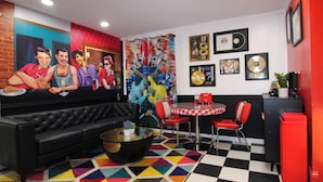 Una habitación con un sofá de cuero negro, un suelo de patrón geométrico y colores vivos, una mesa a cuadros rojos y blancos, y un armario rojo.
