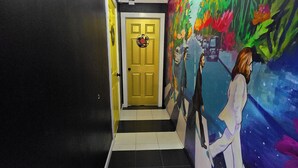 Un pasillo con una puerta amarilla, suelo de baldosas blanco y negro, y un mural colorido en la pared.