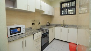 A compact kitchen with white cabinets, a microwave, a stove, and a window with bars. - The Vibrant Loadge (Kigali)