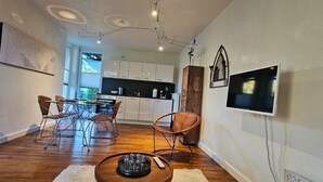Una cocina moderna con armarios blancos, un comedor con mesa de vidrio y sillas de metal, y un televisor montado en la pared.
