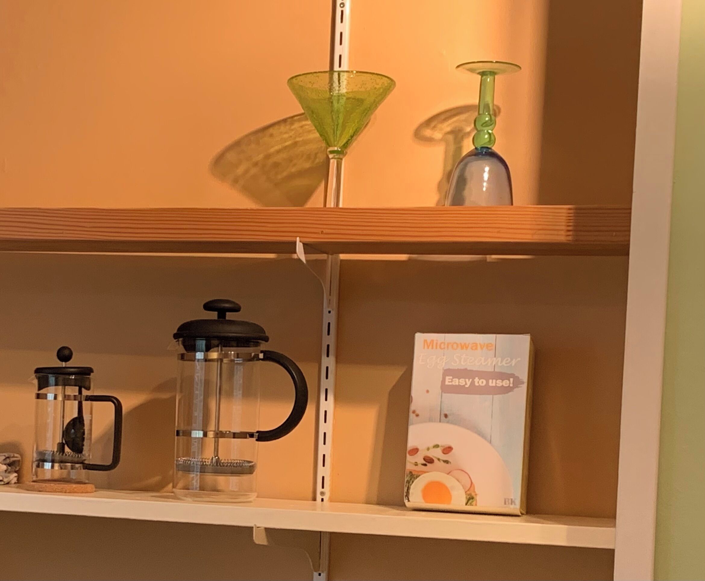 Uma prateleira com uma cafeteira, um copo de vidro verde e a caixa de um cozinhador de ovos elétrico.