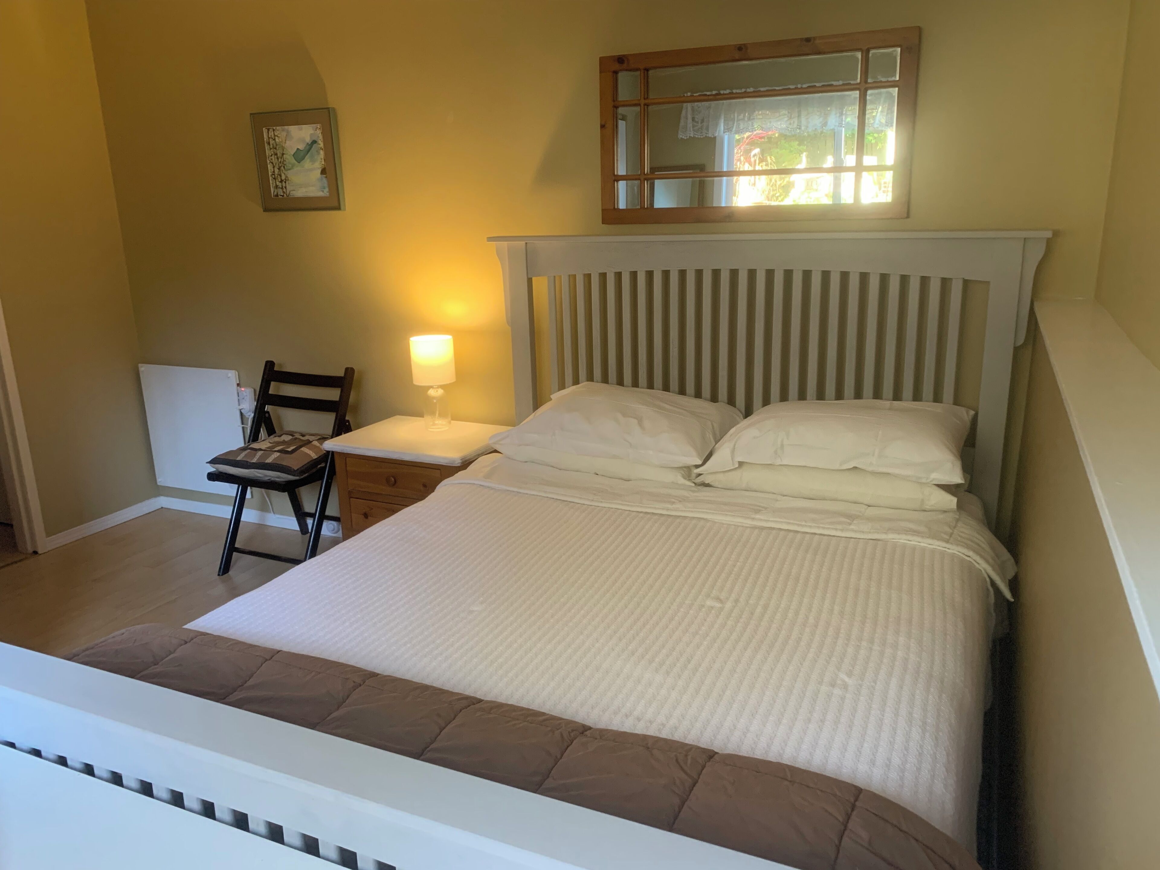 Une chambre bien rangée, avec un lit, une table de chevet dotée d’une lampe, une chaise et une fenêtre donnant sur un paysage verdoyant.