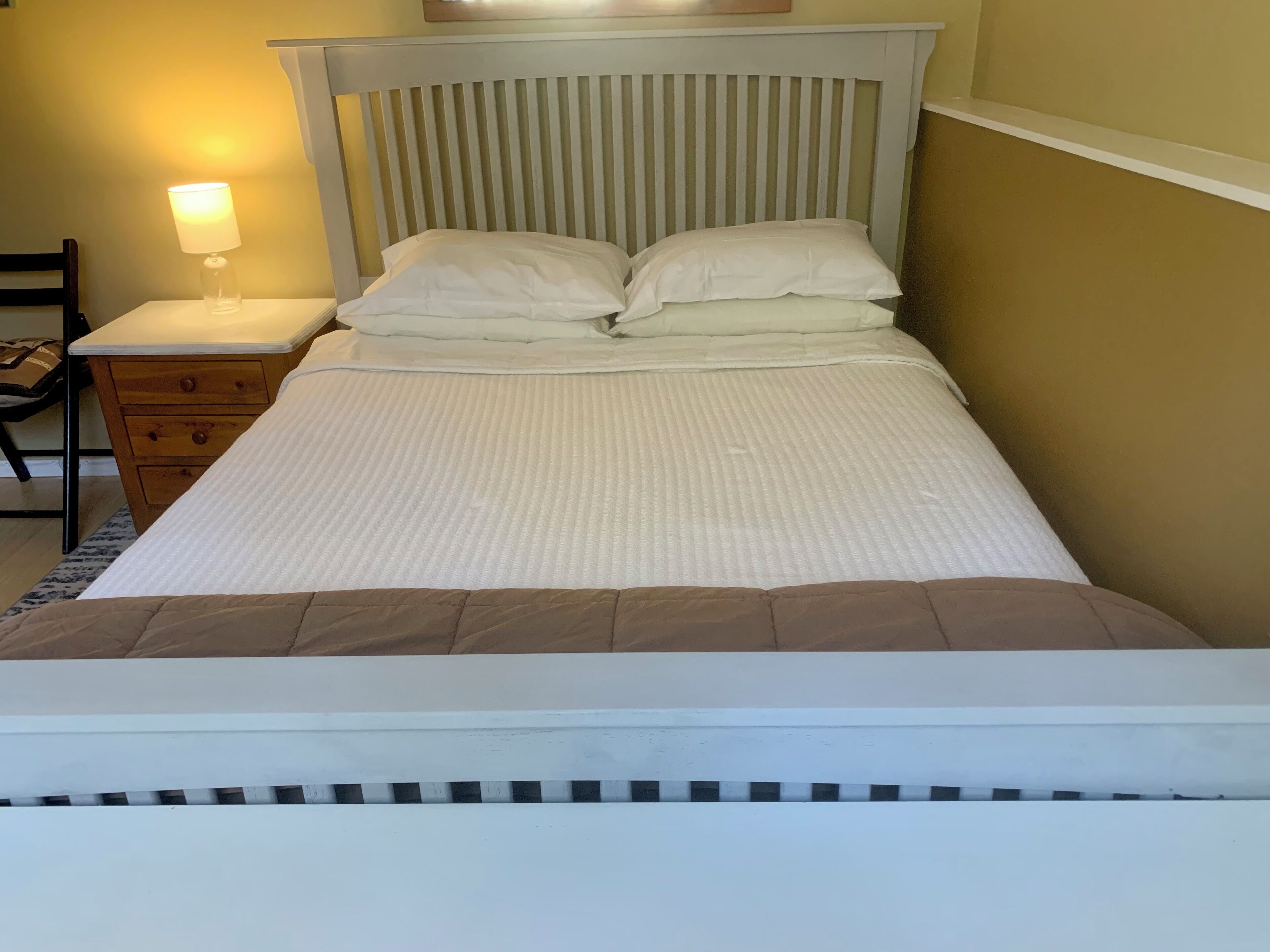 Een net opgemaakt bed met een witte dekbedovertrek en kussens, een houten nachtkastje met een lamp, en een stoel.