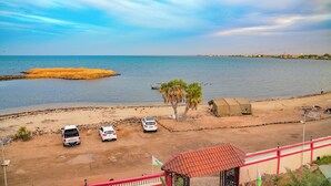 Μια παράκτια περιοχή με αμμώδη παραλία, σταθμευμένα αυτοκίνητα, μια φοινικιά και ένα μικρό κτίριο.