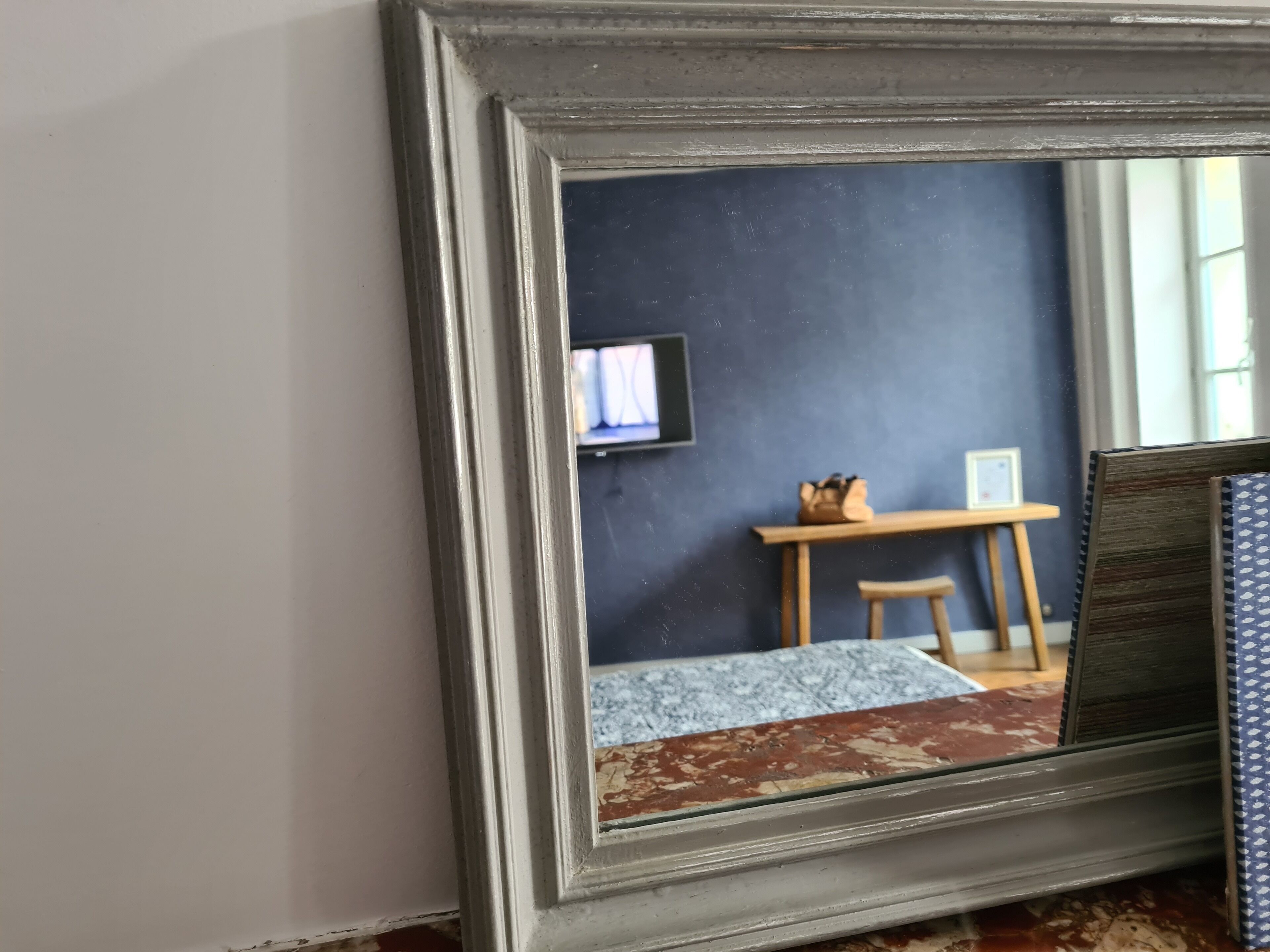 A mirror with a wooden frame reflecting a room with a dark blue wall, a small table, a chair, and a television.