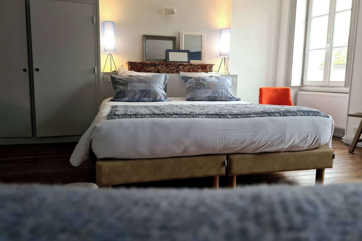 Ein ordentlich gemachtes Bett mit einer gemusterten Decke, zwei Nachttischlampen, einem Spiegel und einem Fenster mit Blick auf Grünflächen.