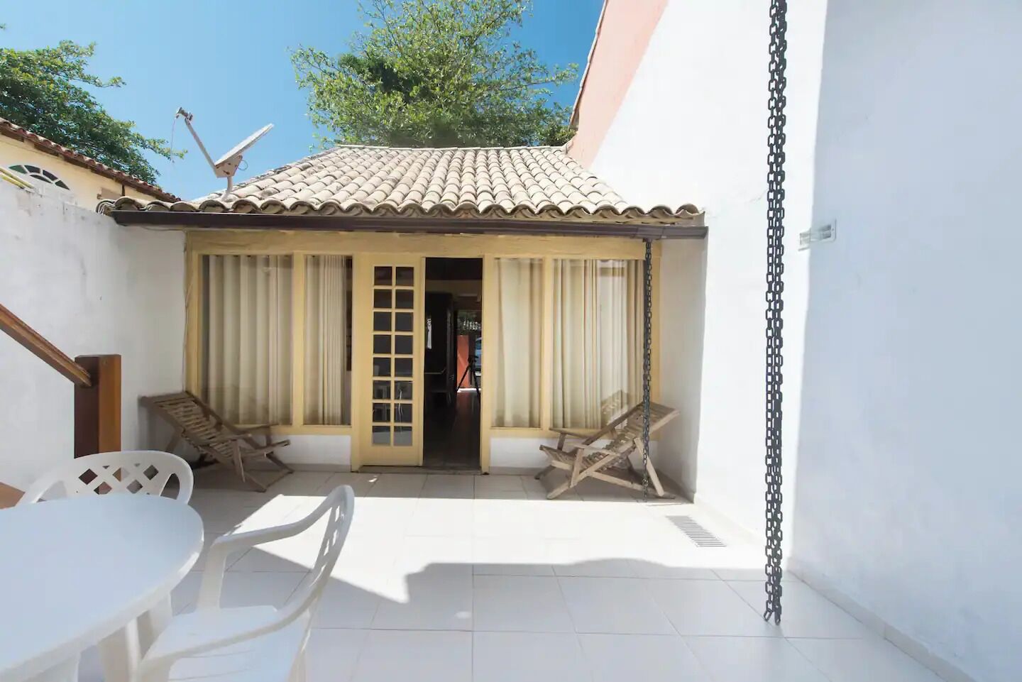 A small patio with a tiled roof, two wooden chairs, a white table, and a door leading to an interior space.