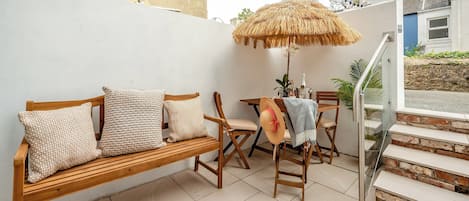 A patio area with a wooden bench, a table with chairs, a thatched umbrella, and a staircase leading to an upper level.
