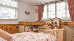 A bedroom with a wooden bed, a dresser, and a vanity with a mirror. There are windows with blinds and curtains, and a small shelf with books and a vase of flowers.