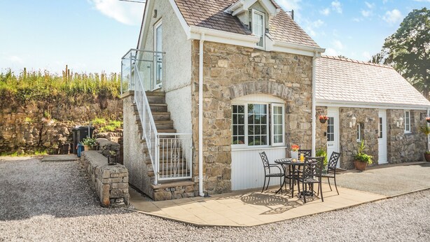 Une maison en pierre avec un toit en pente, un petit balcon et une terrasse aménagée avec du mobilier d’extérieur.