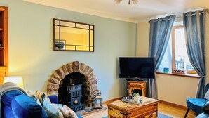 Een woonkamer met een stenen open haard, een televisie op een houten standaard, een blauwe bank en een raam met gordijnen.