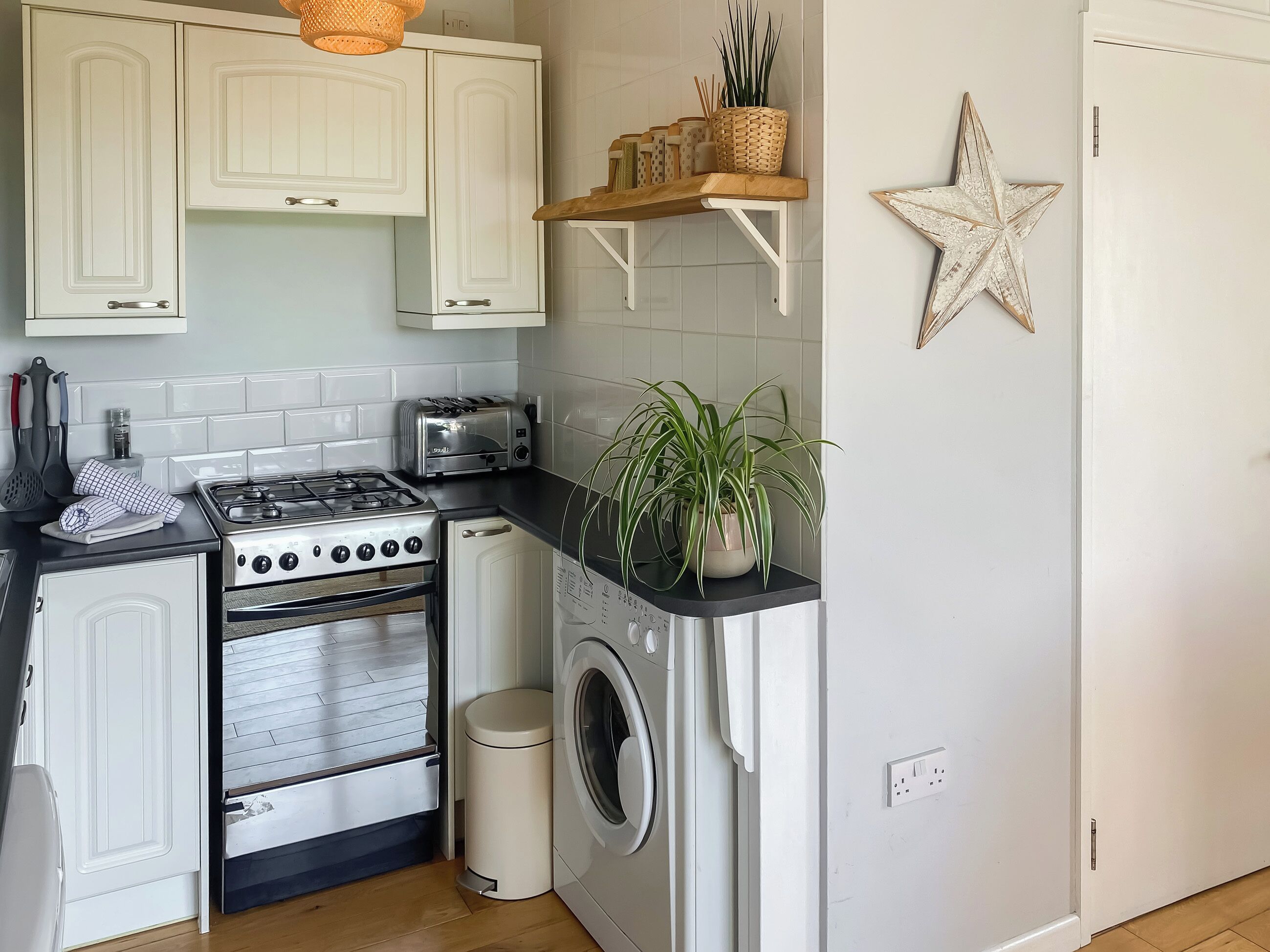 Une cuisine compacte avec des meubles blancs, une cuisinière à gaz, un grille-pain, un lave-linge et une petite poubelle.