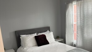 Un dormitorio con cama, mesitas de noche, una ventana con cortinas y una puerta.