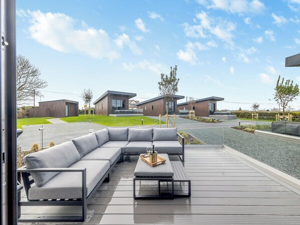 A modern outdoor seating area with a grey sectional sofa and a coffee table, overlooking a residential street with modern houses and a clear sky.