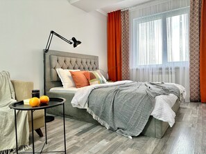 Un dormitorio moderno con cabecera capitonada, una mesita de noche con lámpara y vistas al exterior a través de cortinas traslúcidas.