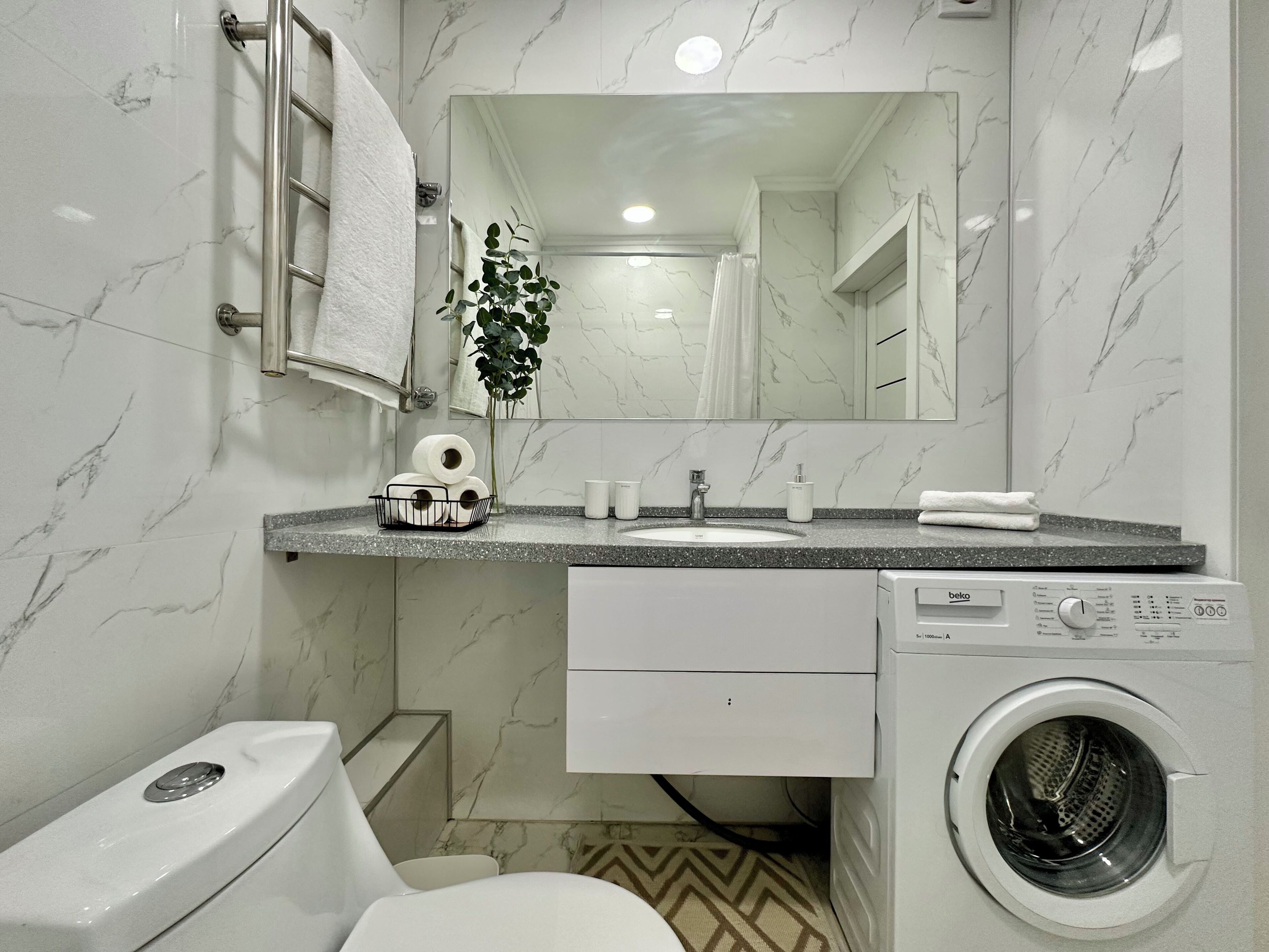 Ein modernes Badezimmer mit Marmor-Spritzschutz, einem Waschbecken mit Spiegel darüber, einer Toilette und einer Frontlader-Waschmaschine.