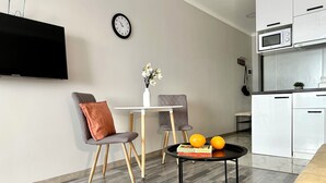 A modern living room with a flat-screen TV, a dining table with chairs, a small side table with books and oranges, and a kitchen area with a microwave.