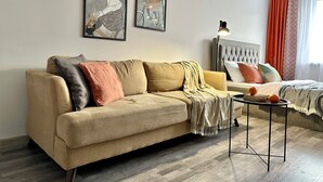 A modern living room with a beige sofa, a small coffee table, and a bed with a tufted headboard. There are two framed artworks on the wall and a lamp on the side table.