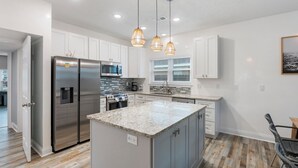 A modern kitchen with white cabinets, a stainless steel refrigerator, a granite countertop, and pendant lighting. - PCB Paradise Sands Oasis - Private Pool, Game Room, Hot Tub! (Panama City Beach)