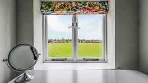 Une fenêtre ornée d’une lambrequin à motifs floraux, un miroir posé sur une surface blanche et une vue sur un champ verdoyant.