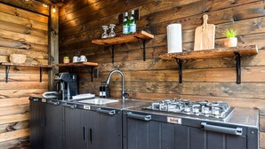 A compact kitchen with a built-in gas stove, a sink, and wooden shelves with kitchenware. - The Tree Spa, Tiny Cabin w/ Hot Tub in the trees (Hayesville)