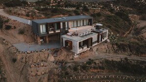 A modern two-story house with a flat roof, large windows, and a patio area with outdoor seating. - Kasa Kefi, Luxurious Vineyard Oasis in Kasa Kava Valle d Guadalupe (Valle de Guadalupe)