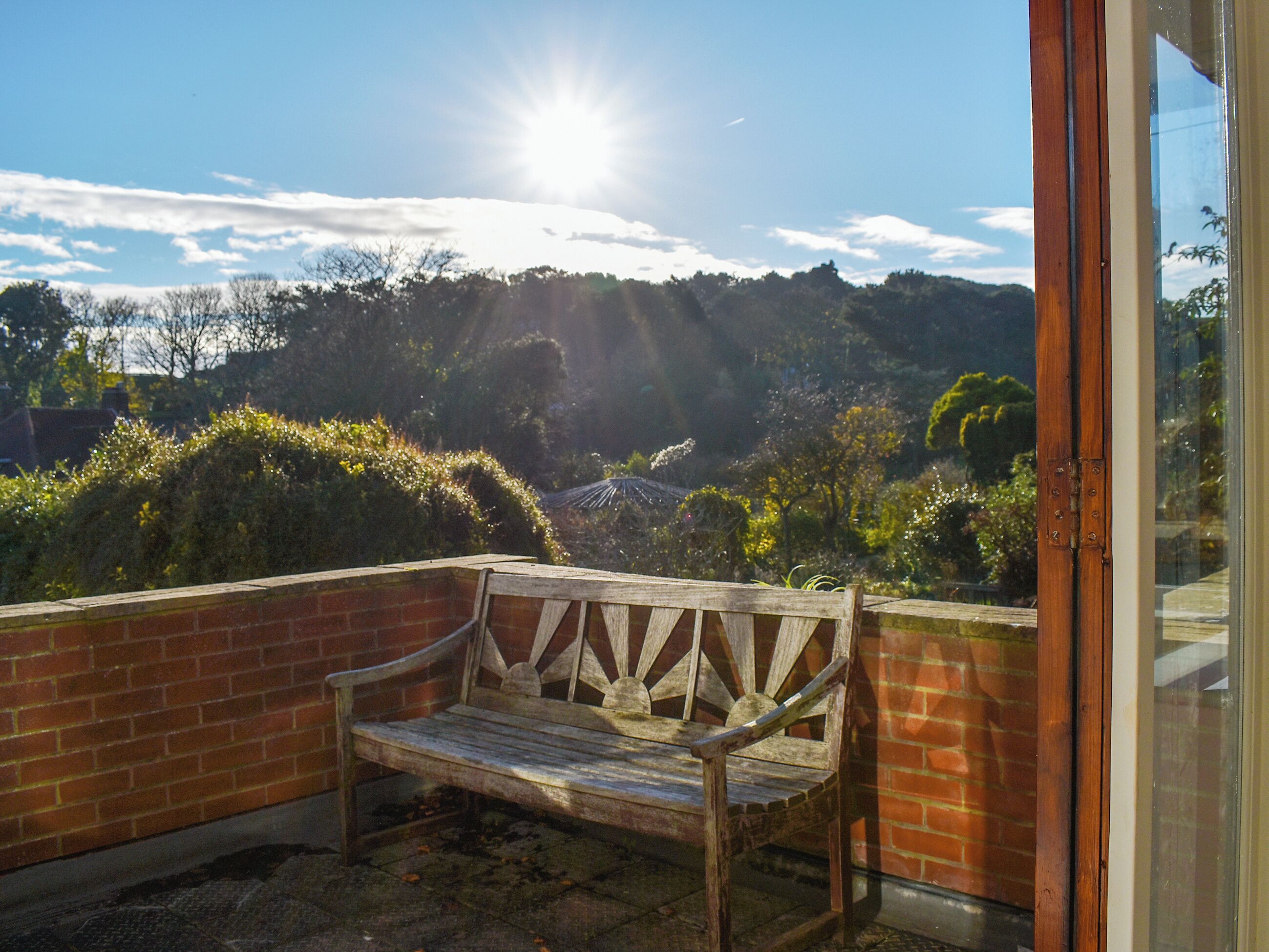 Un banc en bois se trouve à l’extérieur d’un bâtiment doté d’une porte en verre, et offre une vue sur un paysage vallonné, avec des arbres et un ciel dégagé.