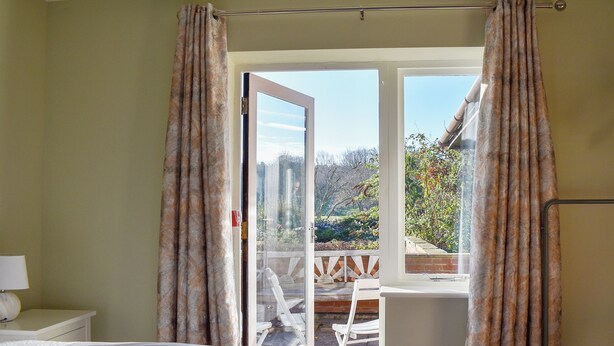 A bedroom with a bed, a window with curtains, and a view of a garden and outdoor seating area. - Pines Cottage - UK42110 (St. Margaret&rsquo;s Bay)