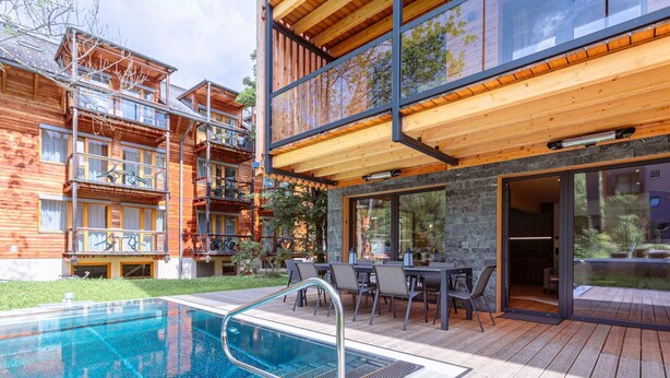 A modern outdoor pool area with a wooden deck, a glass-enclosed dining area, and a view of a multi-story building with balconies. - Appealing Apartment With Pool (Sankt Georgen am Kreischberg)