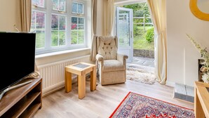 Un salon chaleureux avec un téléviseur, une petite table, un fauteuil confortable et une vue sur un jardin, visible à travers une porte vitrée.