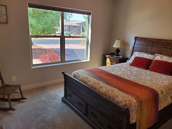 A bedroom with a large window, a bed with a patterned bedspread, a wooden nightstand, and a chair.