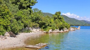 Visão nítida de uma praia de seixos, com mar calmo, rodeada por árvores verdes densas e afloramentos rochosos.