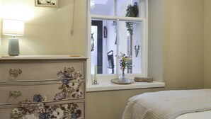 A bedroom with a bed, dresser with floral drawers, a window with a view, and a bedside lamp.