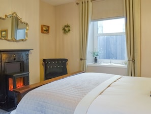 Ein Schlafzimmer mit Bett, Kamin, Sessel, einem Fenster mit Vorhängen und einem Spiegel an der Wand.