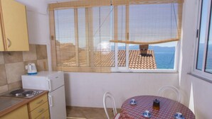 A compact kitchen with a view of the sea, a small dining table with a checkered tablecloth, and a white refrigerator. - Triple Room with Private Balcony (Brist)