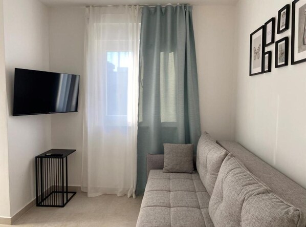 A living room with a grey sofa, a black TV on the wall, a small black side table, and a window with sheer curtains.