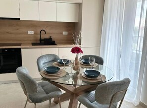 A modern kitchen with white cabinets, a black oven, and a glass dining table with four grey chairs.