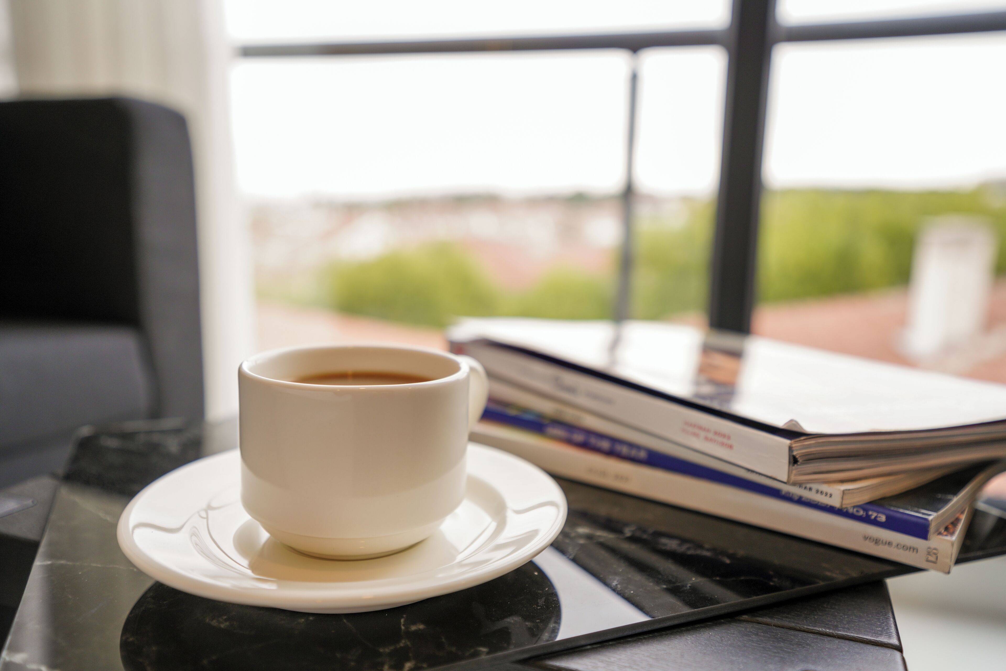 Una taza de café sobre un plato, una pila de revistas y una tableta sobre una mesa cerca de una ventana.