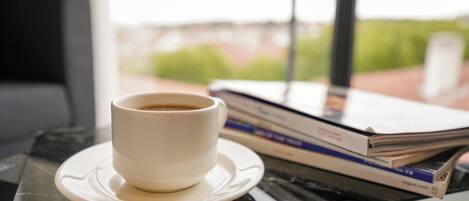 Una taza de café sobre un plato, una pila de revistas y una tableta sobre una mesa cerca de una ventana.