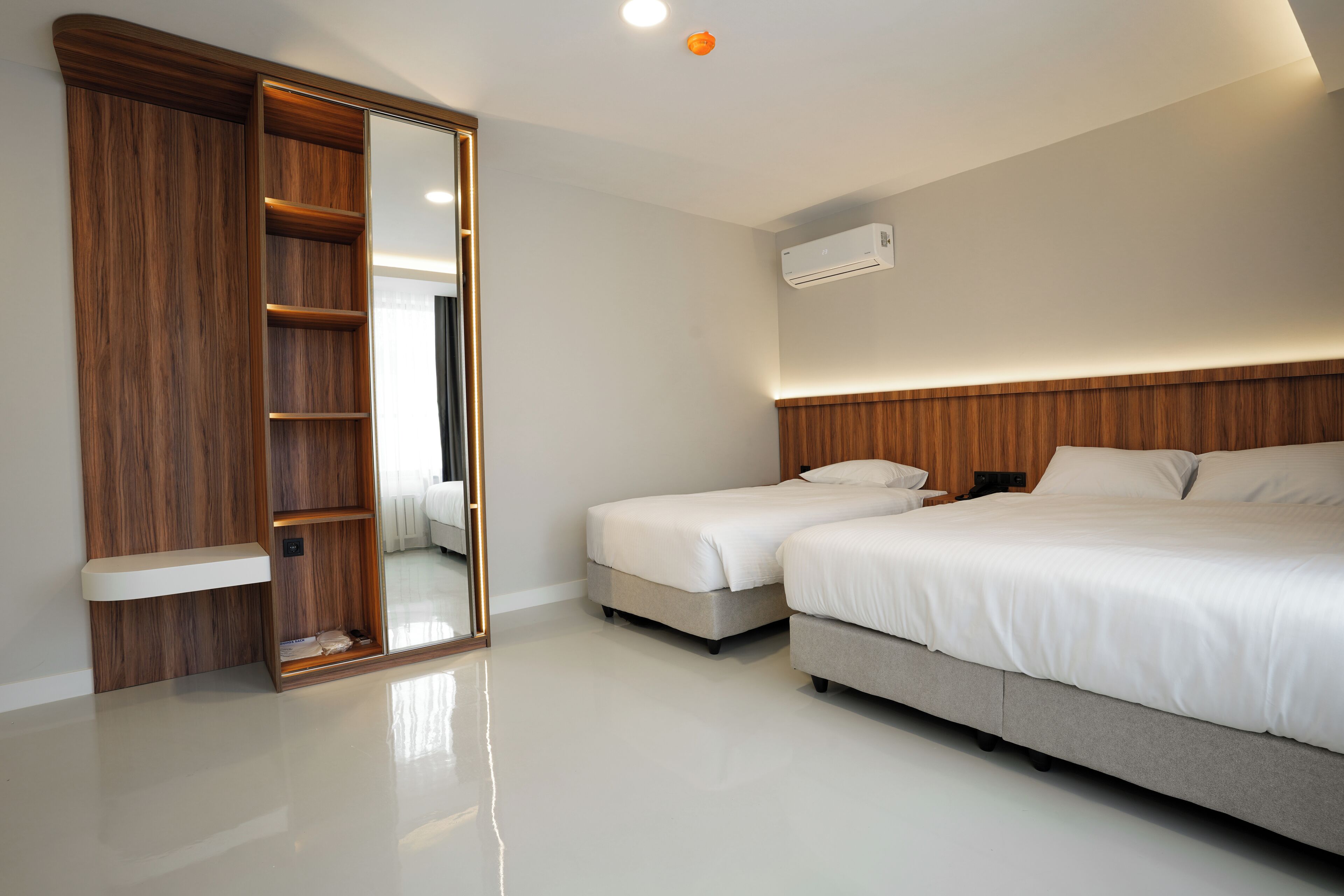 A modern bedroom with a wooden wardrobe, a large mirror, a bed with white bedding, and a ceiling-mounted air conditioning unit.