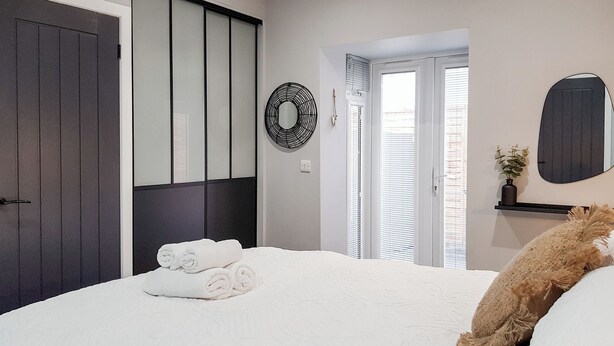A bedroom with a bed, a dark door, a window with blinds, a wall-mounted mirror, and a small shelf with a plant. - 1 bedroom accommodation in Anstruther (Anstruther)