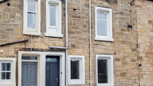 A two-story building with a stone facade, multiple windows, and a door with a number plate. - 1 bedroom accommodation in Anstruther (Anstruther)